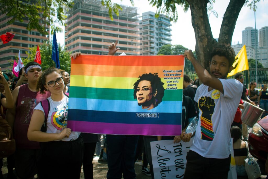 Colorful pride parade featuring diverse crowd holding a statement banner on city streets.