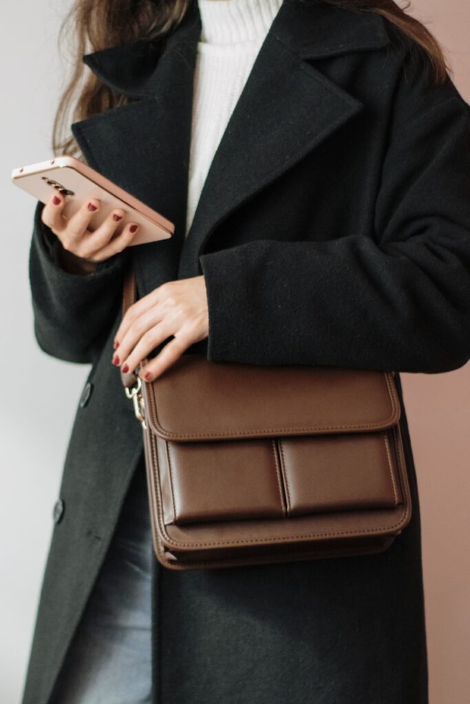 Close-up of a person holding a smartphone, wearing a black coat and brown leather bag, highlighting fashion style.